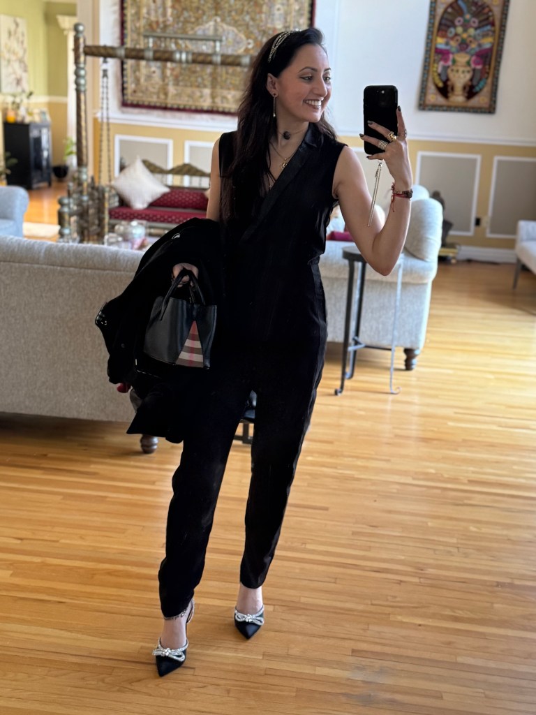 A woman wearing a black tuxedo jumpsuit with satin lapels, accessorized with diamond and gold jewelry, a bejeweled headband, and Swarovski-studded heels, stands in a stylish interior while holding a black purse.